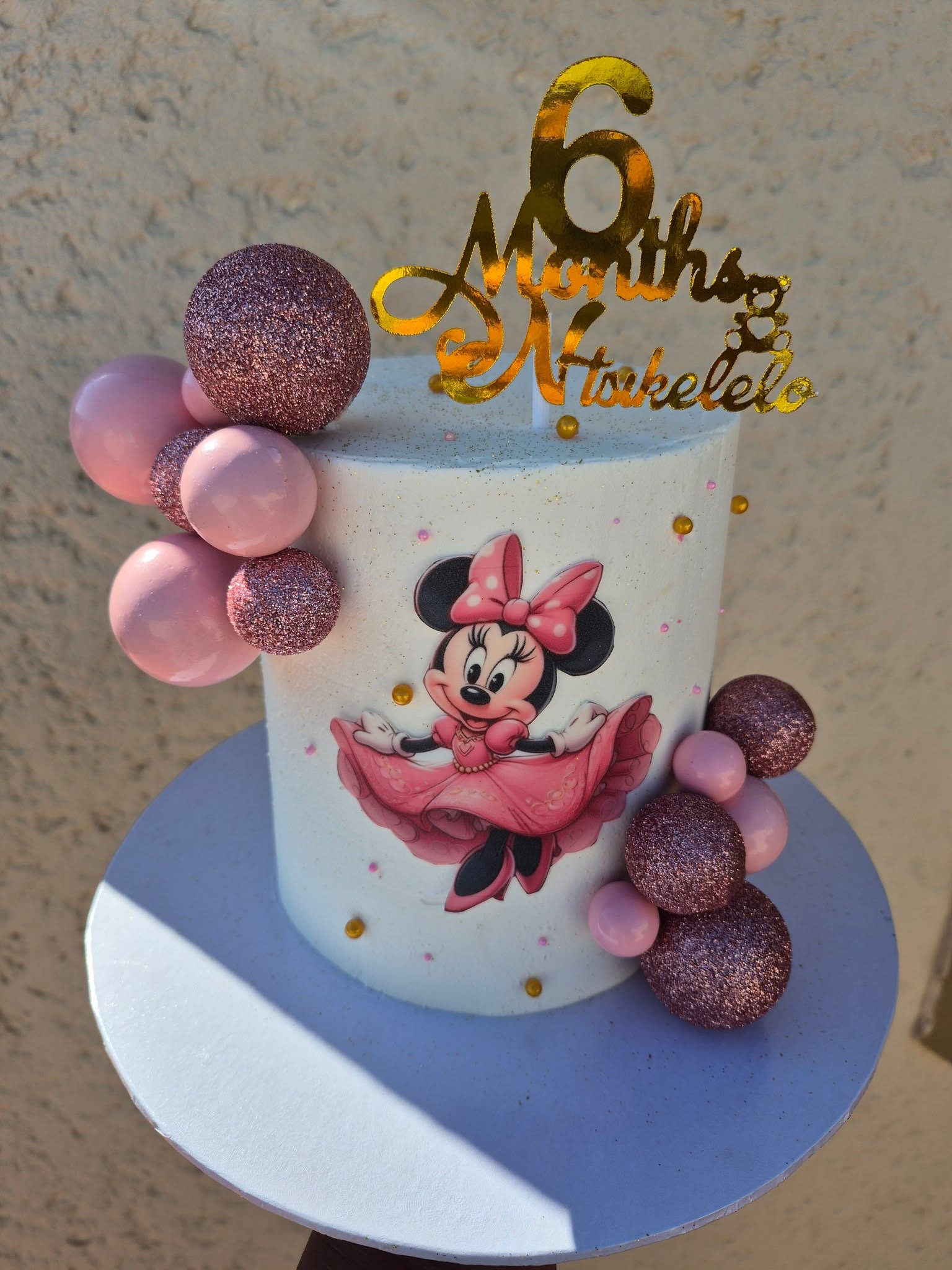A white cake decorated with a Minnie Mouse image and rose gold sphere accents, made by Rosy's Cakes and Events